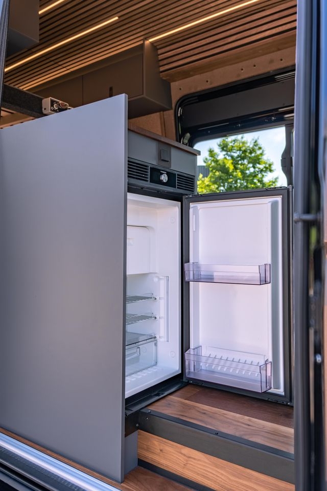 geöffneter Kühlschrank. Eingebaut in ein graues Küchenmöbel in einem Camper Van. Der Boden ist aus Parket. Wie der Rest des Ausbaus von Cocoon Vans ist der Boden aus amerikanischen Nussbaum. Die Beleuchtung an der Decke ist eingeschalten.