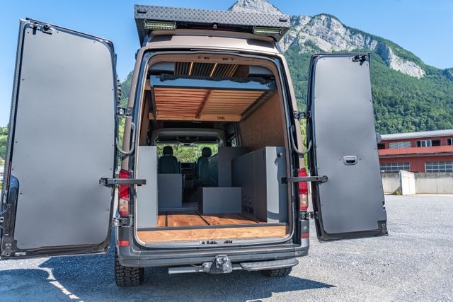 Ein Blick in den VW Crafter 4x4 mit geöffneten Hecktüren. Der Camper Van von Cocoon Vans verfügt über ein Hubbett, welches auf dem Bild oben ist. Man hat einen guten Blick in den ganzen Camper Van. Der Innenausbau ist aus amerikanischem Nussbaum gefertigt. In den Boden sind Airlineschienen für die Ladesicherung eingelassen. Seitlich sind graue Möbel eingebaut, in denen die ganze Technik verstaut ist.